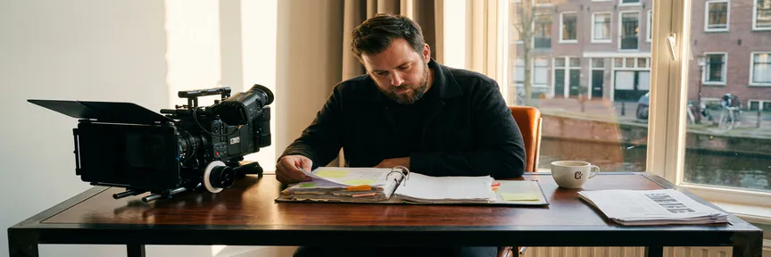 Videodirecteur bestudeert productieplanningmap aan Amsterdams studiobureaus met camera en shotlijsten in warm amberkleurig licht.