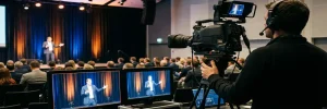 Cameraman filmt zakelijk evenement met spreker op verlicht podium, broadcast monitors op voorgrond, marineblauw en amber verlichting.