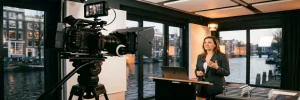 Professionele videocamera op statief filmt een presenterende zakenvrouw in een Amsterdams studio met warm amber licht.