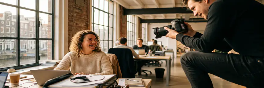 Fotograaf op ooghoogte die lachende collega vastlegt op kantoor in Amsterdam, warme middaglichtval door industriële ramen.