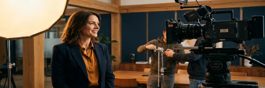Zelfverzekerde professional voor een filmcamera op een modern Amsterdams kantoorset, warm sleutellicht, crew op de achtergrond.