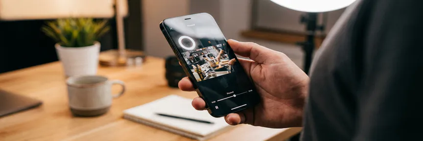 Smartphone verticaal vastgehouden boven een Amsterdams studiobureaus, scherm verlicht met een bevroren korte videoclip, ringlight zacht gereflecteerd.