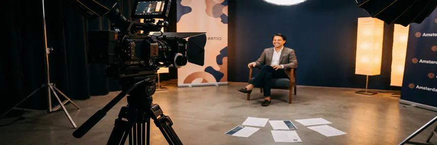 Professionele videocamera op statief gericht op sollicitant in modern Amsterdams studio met softboxverlichting en cv's op betonnen vloer.